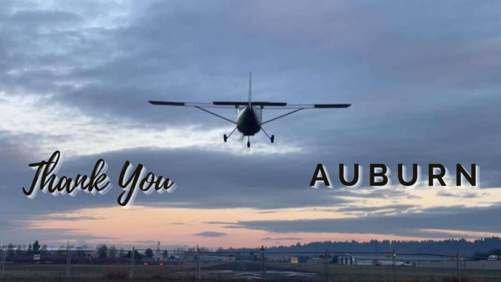 A prop plane lands at Auburn Municipal Airport in Auburn WA with the sunrise in the background. The words Thank You Auburn are written on the image.
