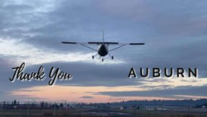 A prop plane lands at Auburn Municipal Airport in Auburn WA with the sunrise in the background. The words Thank You Auburn are written on the image.